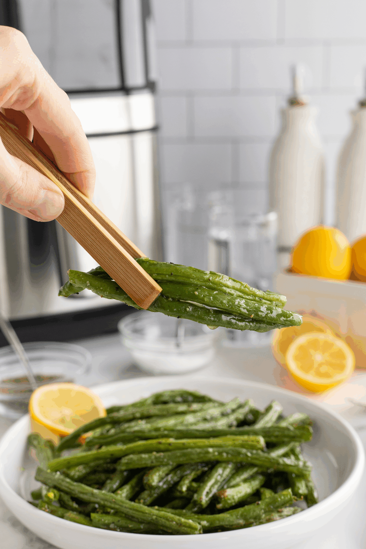 A hand uses wooden tongs to pick up seasoned green beans from a white bowl, with an air fryer, cut lemons, glassware, and bottles visible in the background on a kitchen counter.