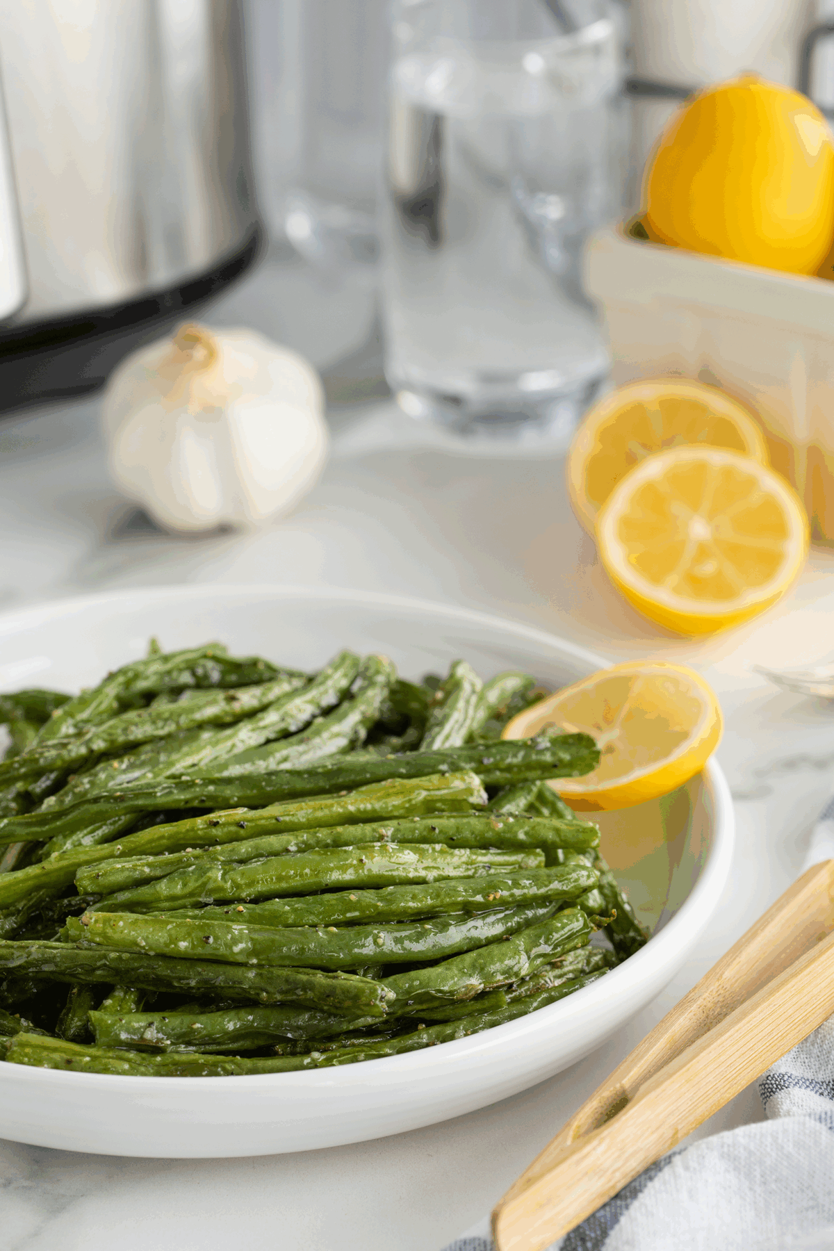 A white plate filled with cooked green beans garnished with a lemon slice. In the background, there are fresh lemons, a garlic bulb, a glass of water, and kitchen items on a white countertop.