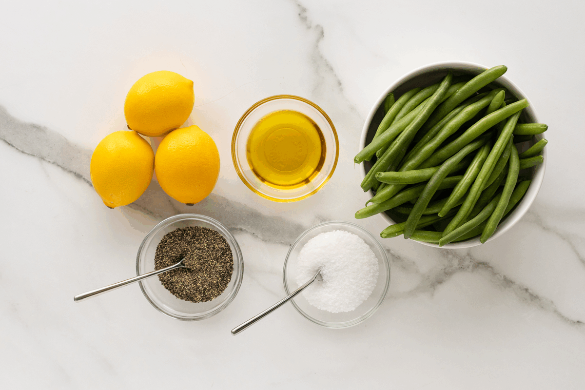 Three lemons, a bowl of fresh green beans, a small bowl of olive oil, ground black pepper, and coarse salt in glass bowls with spoons, all arranged on a white marble surface.