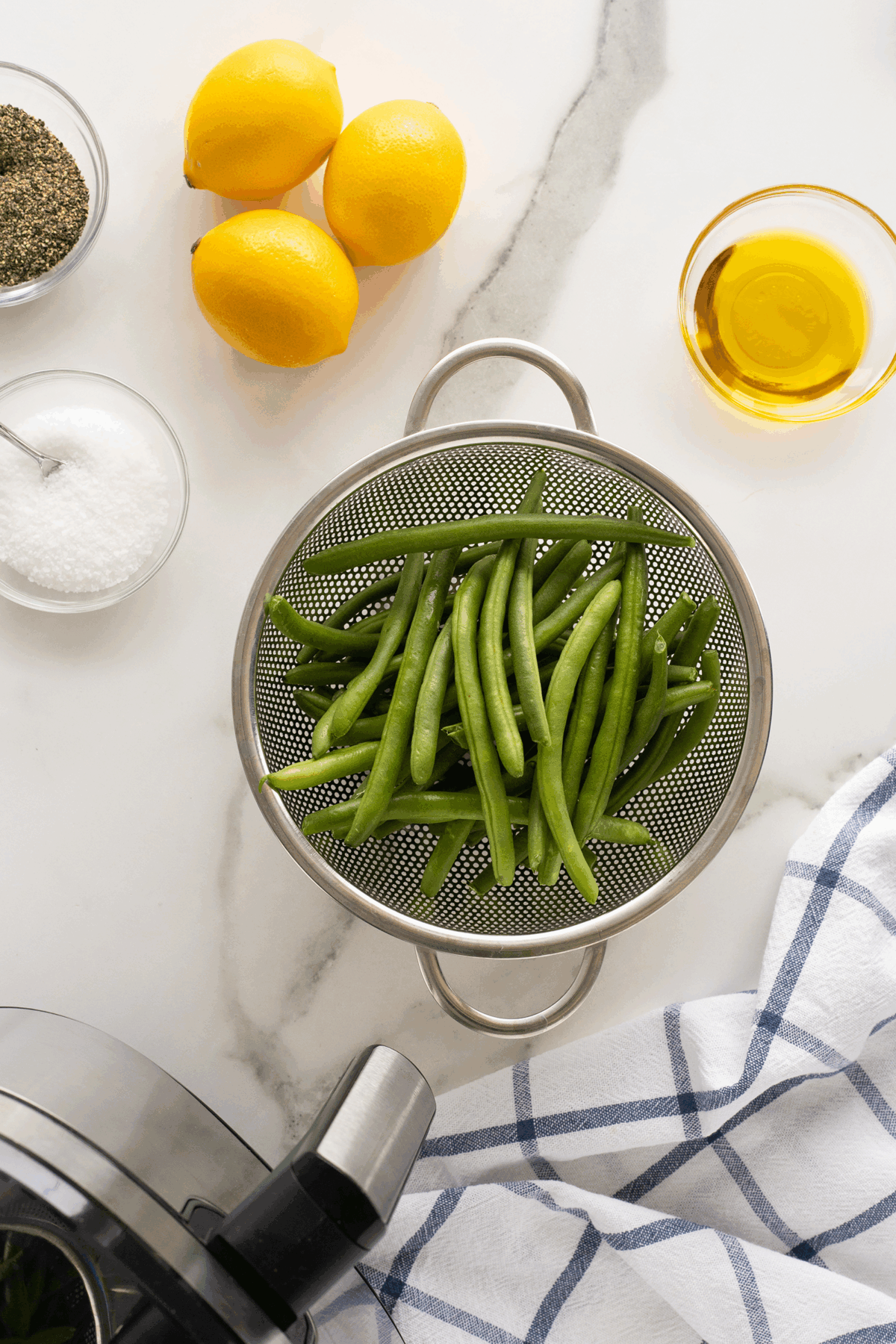 Overhead view of fresh green beans in a strainer, with two lemons, a bowl of olive oil, a bowl of salt, a bowl of pepper, and a blue-and-white striped towel on a marble countertop.