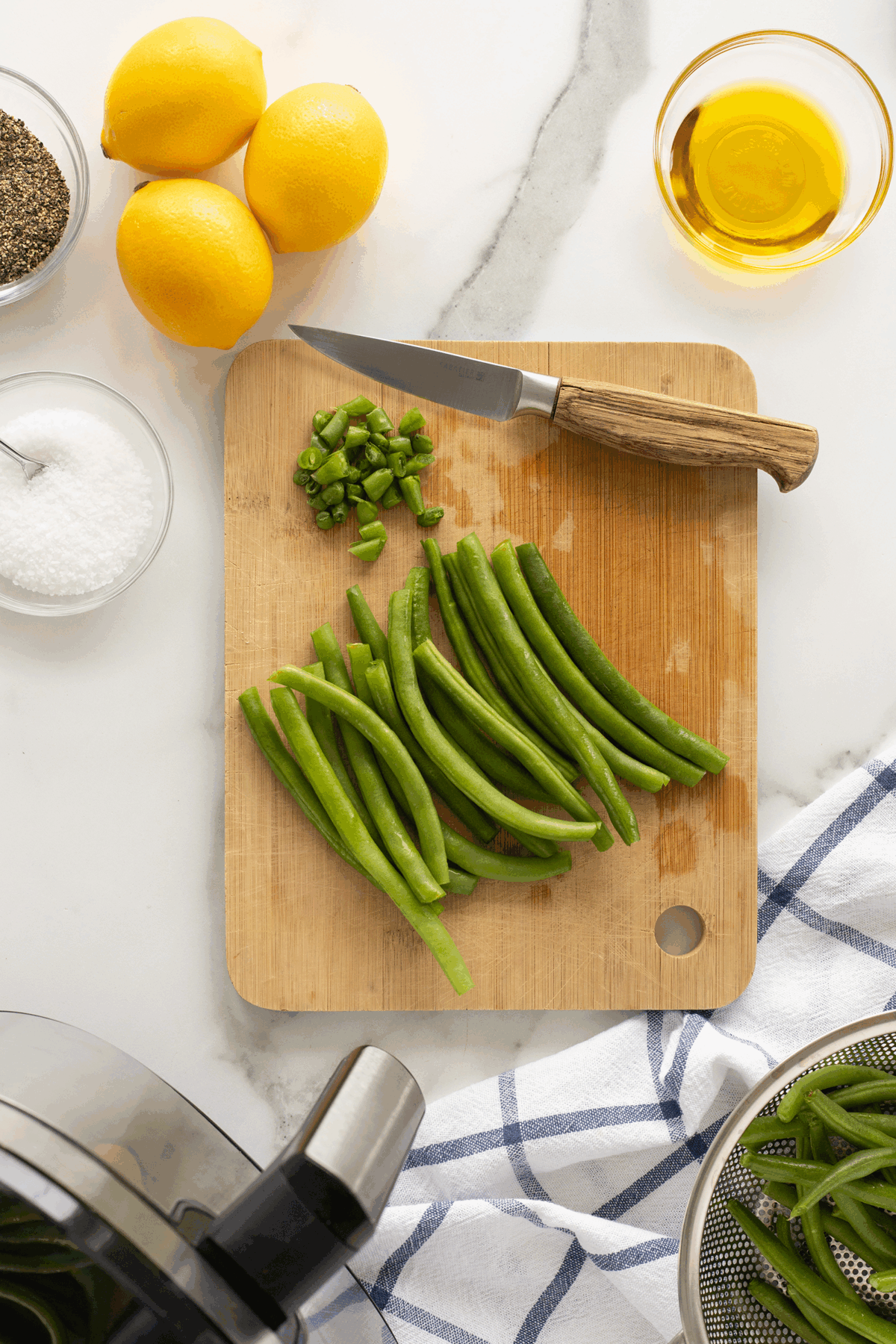 A wooden cutting board with fresh green beans, chopped green pepper, and a paring knife. Nearby are lemons, olive oil, salt, pepper, a striped towel, and an air fryer basket partially visible in the corner.