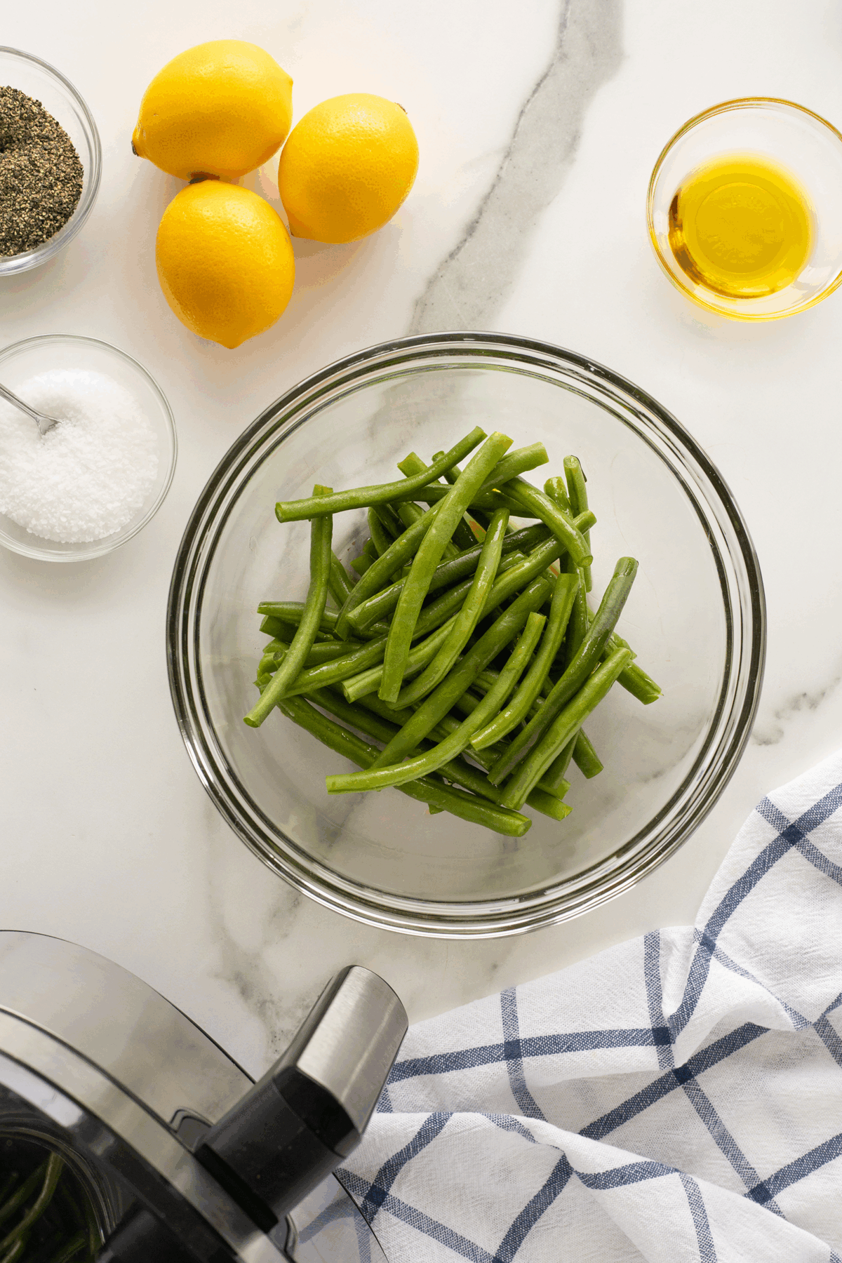 A glass bowl filled with fresh green beans sits on a marble countertop, surrounded by three lemons, a bowl of salt, a small dish of olive oil, ground pepper, and part of an air fryer with a blue striped towel nearby.