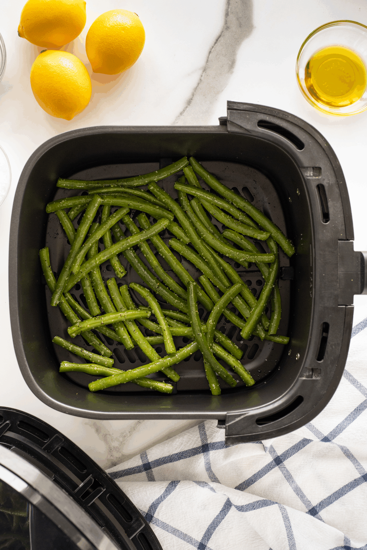 Green beans seasoned with olive oil are arranged in the basket of an air fryer. Fresh lemons, a bowl of olive oil, and a blue-and-white towel are nearby on a marble countertop.