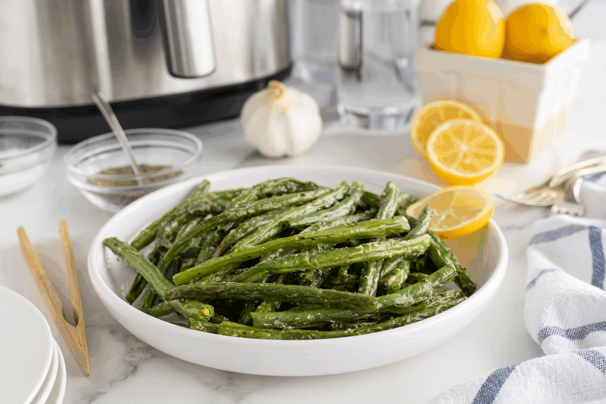 A white bowl filled with cooked green beans seasoned with herbs, placed on a white countertop. Nearby are lemons, a garlic bulb, spices in small bowls, a fork, and a striped napkin.