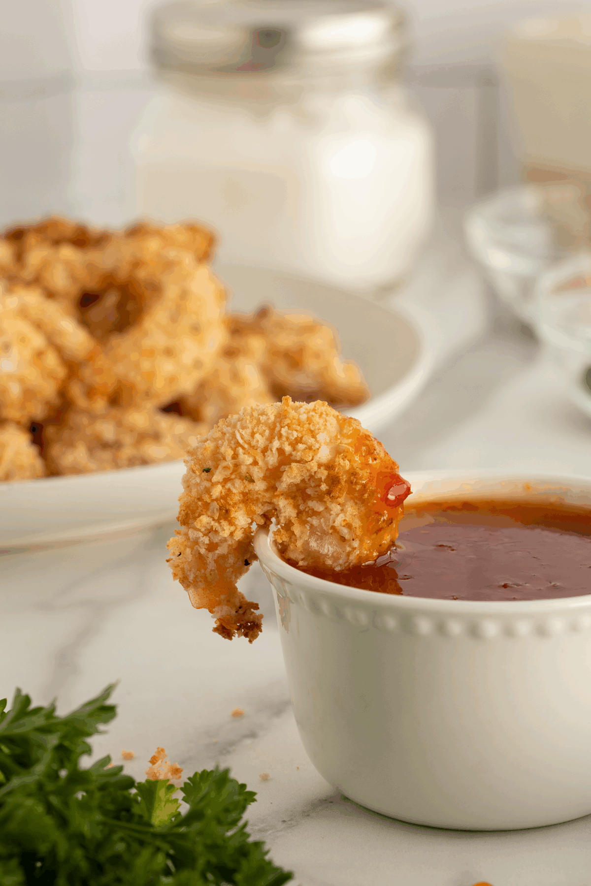 A breaded shrimp is being dipped into a cup of red sauce, with a plate of more breaded shrimp and a jar in the background. Fresh parsley sits in the foreground on a marble surface.