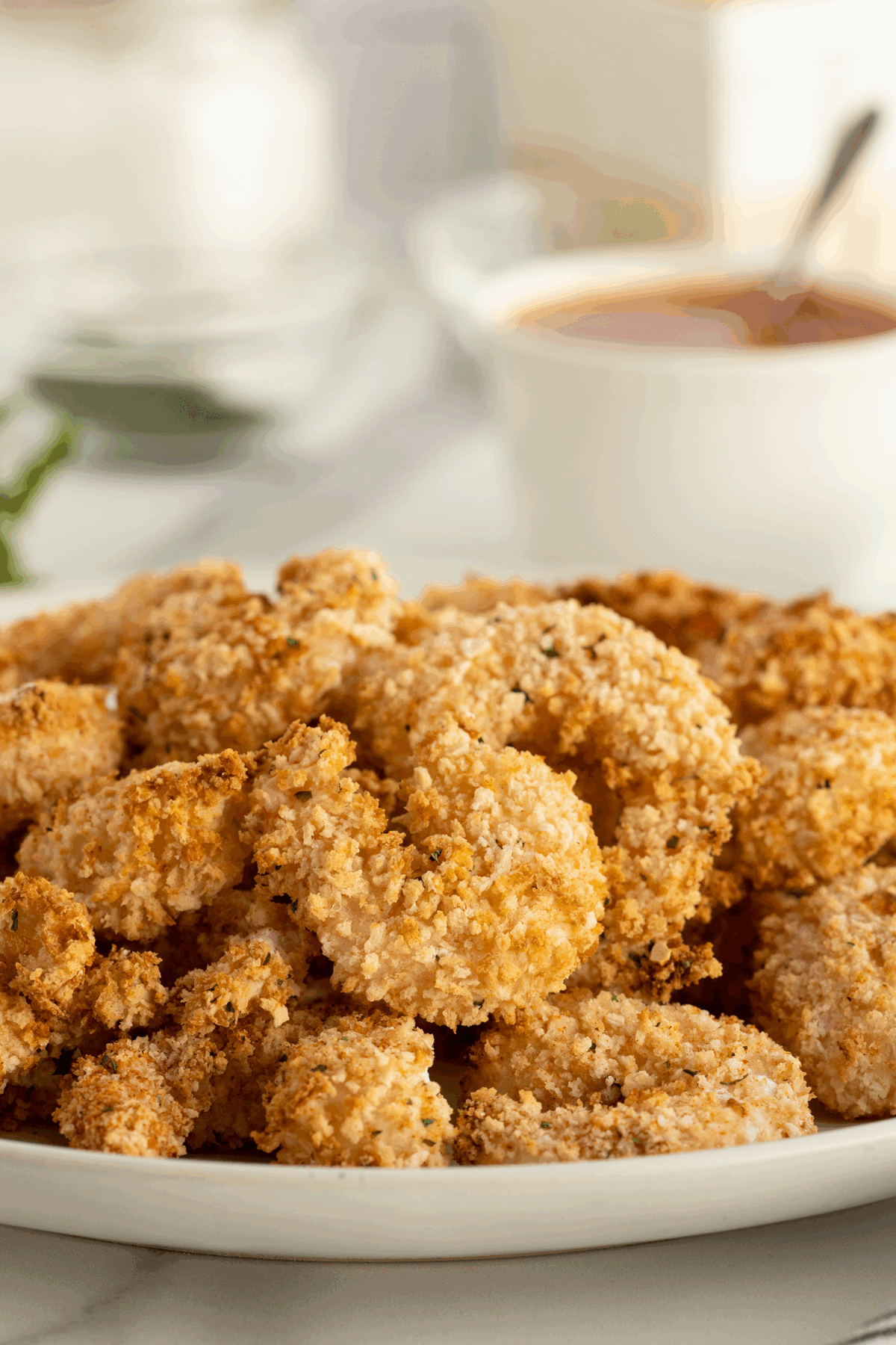 A plate piled with golden, crispy breaded shrimp, with a bowl of dipping sauce and blurred kitchen items in the background.