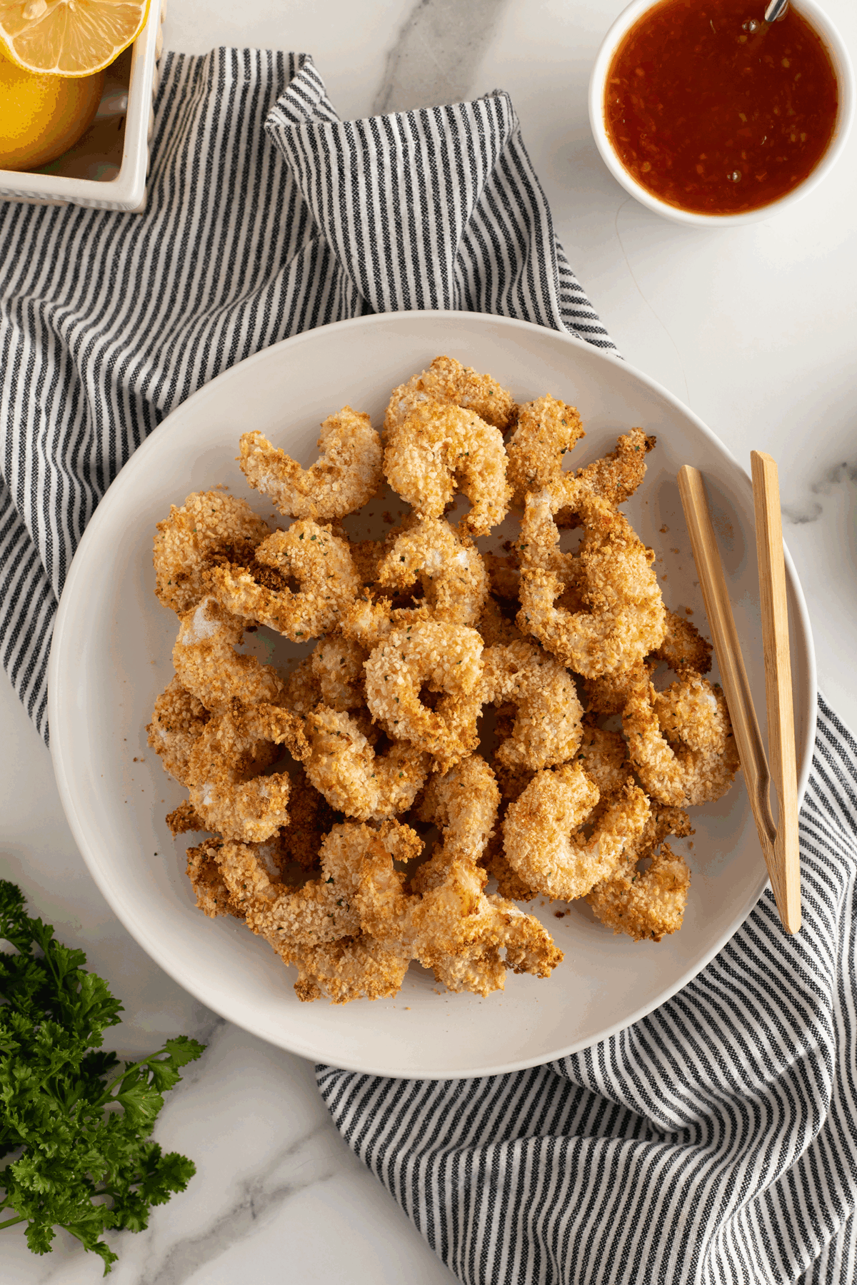 A white plate of breaded, baked shrimp with chopsticks on a striped cloth, next to a bowl of red dipping sauce, lemon wedge, and parsley on a marble surface.