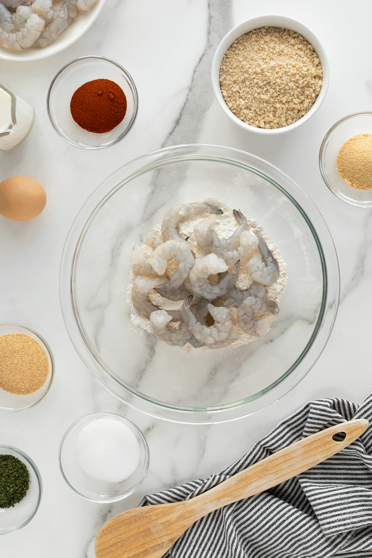 A glass bowl of raw shrimp surrounded by bowls of breadcrumbs, seasonings, an egg, and a wooden spoon on a white marble surface with a striped towel.