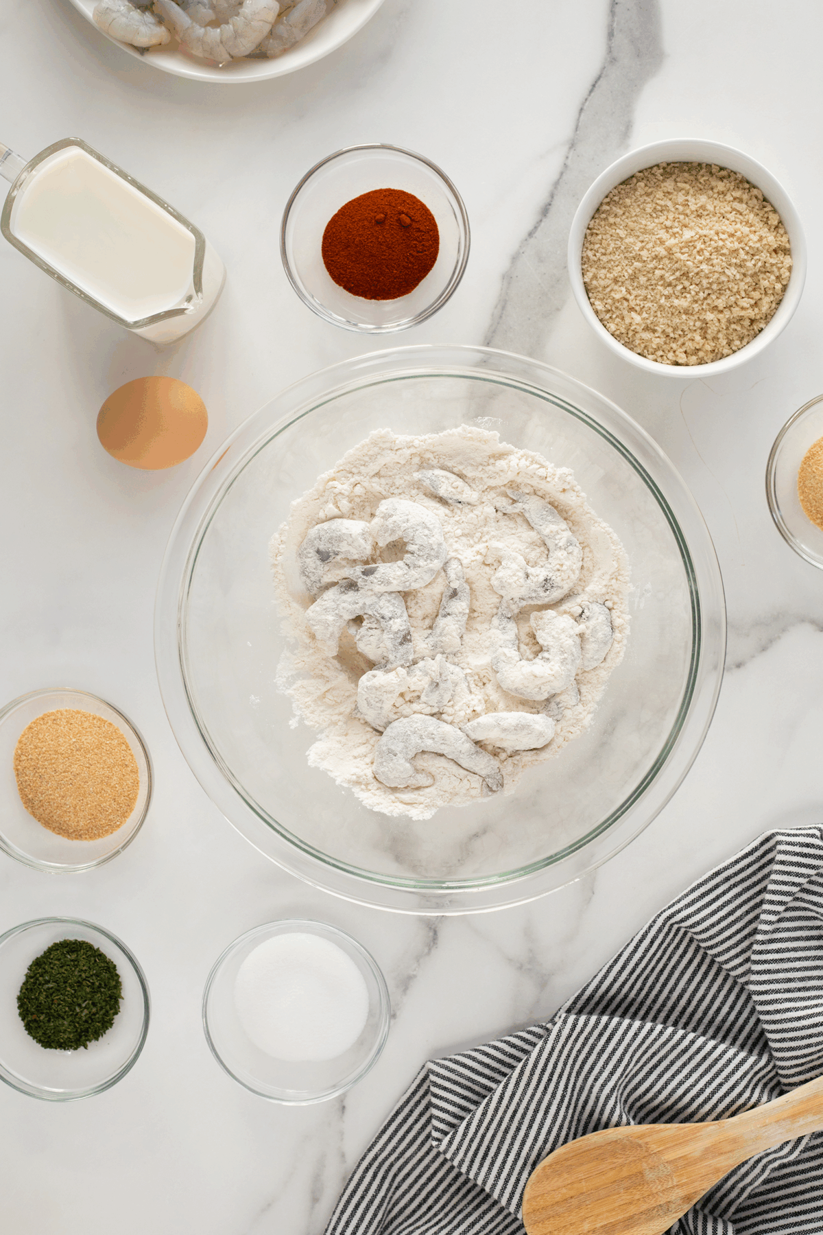 A glass bowl with shrimp coated in flour sits on a marble countertop, surrounded by milk, paprika, panko breadcrumbs, an egg, sugar, spices, and a striped towel with a wooden spoon.