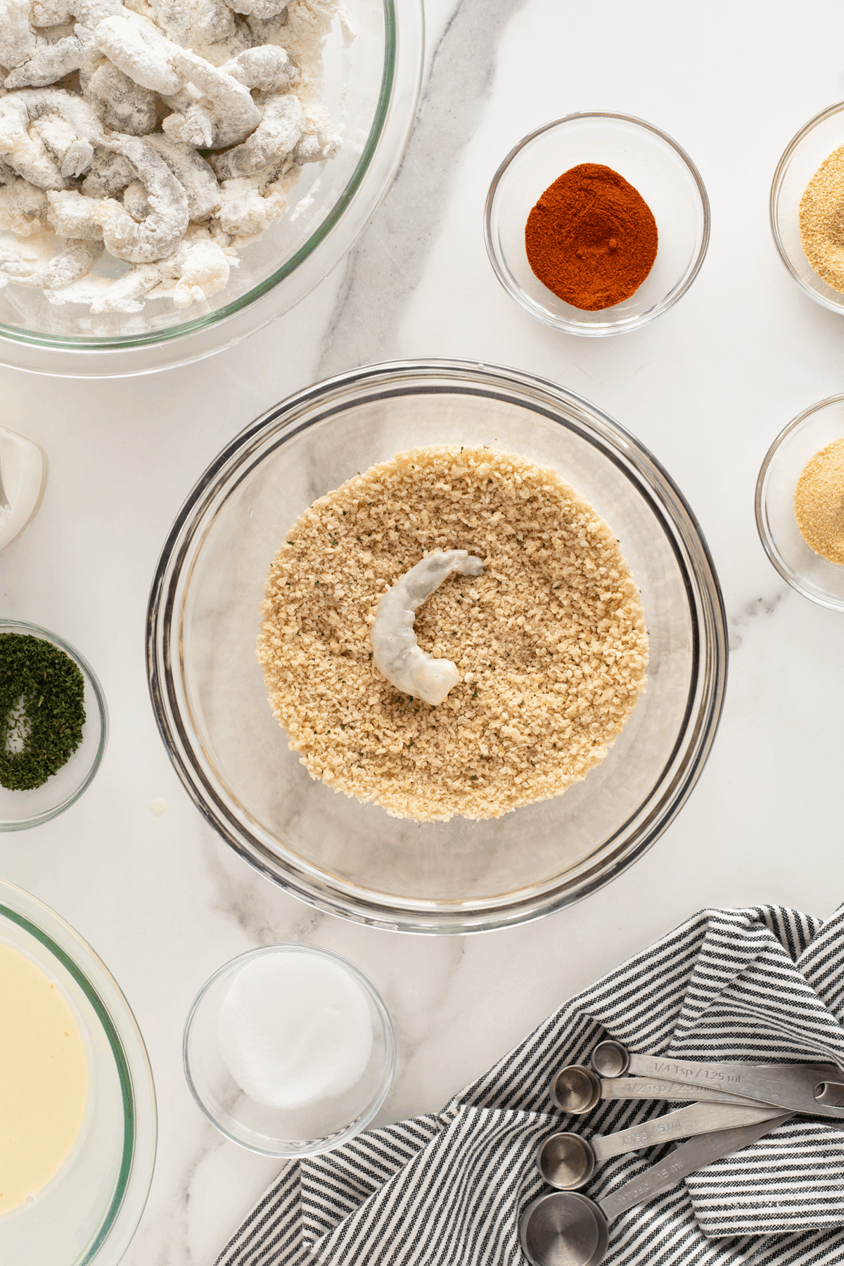 A breaded shrimp is in a glass bowl of breadcrumbs, surrounded by bowls of spices, batter, and coated shrimp on a white marble surface, with a striped cloth and measuring spoons nearby.