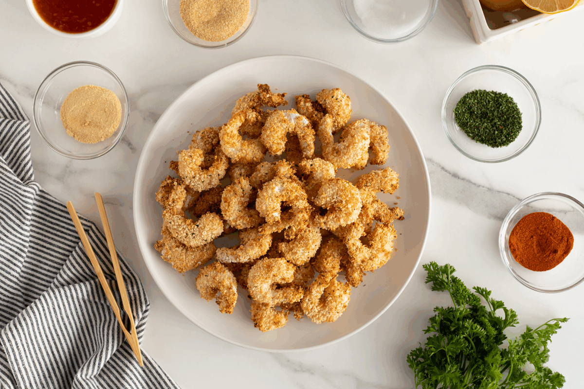 A plate of breaded, baked shrimp is surrounded by small bowls of various seasonings and herbs on a marble countertop, with a striped towel, parsley, and chopsticks nearby.