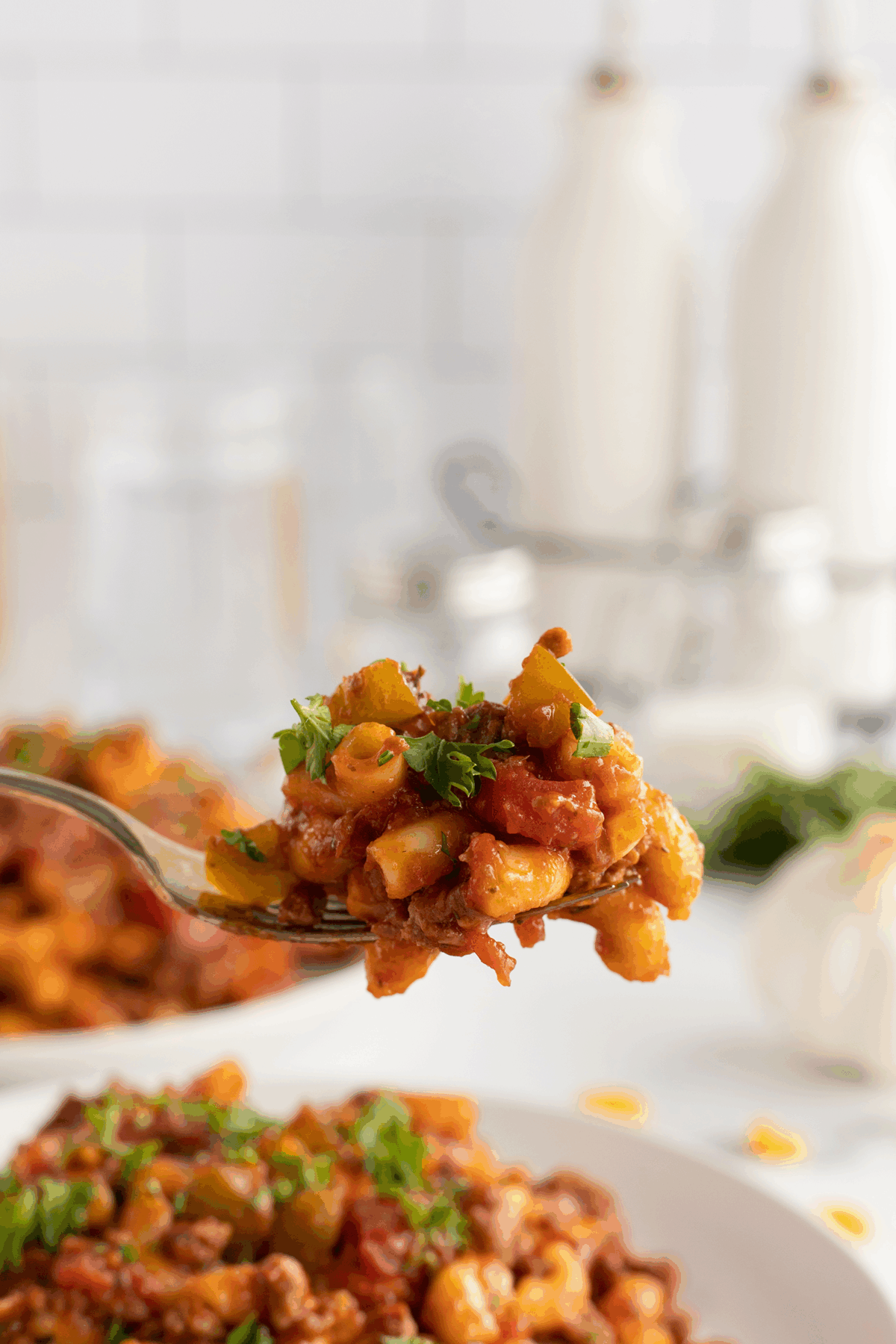 A close-up of a fork holding a bite of pasta with tomato sauce, ground meat, and herbs. In the background, a plate of the same dish sits on a white table with blurred kitchen items.