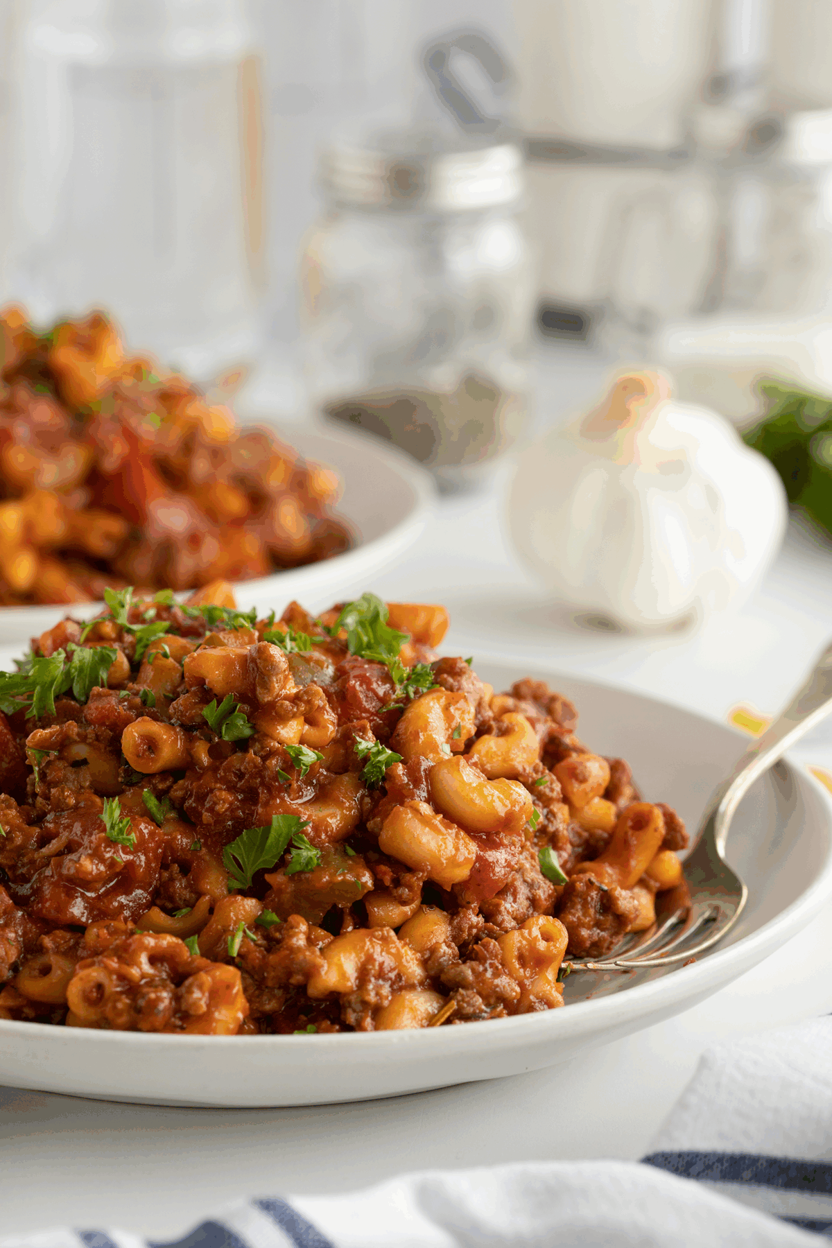 A close-up of a plate of macaroni pasta with ground beef, tomato sauce, and garnished with fresh parsley. A fork rests on the plate; garlic and seasonings are blurred in the background.