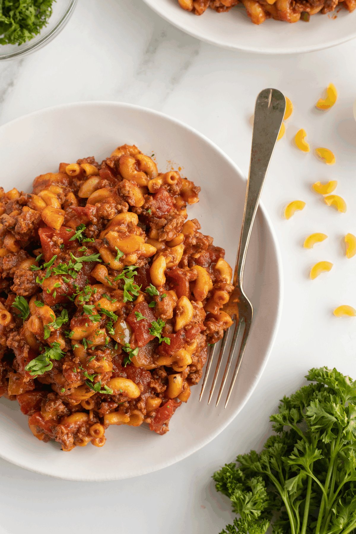 A white plate of seasoned macaroni with ground beef, tomato sauce, and herbs, garnished with chopped parsley. A fork rests on the plate, with parsley and uncooked macaroni scattered nearby on a white surface.