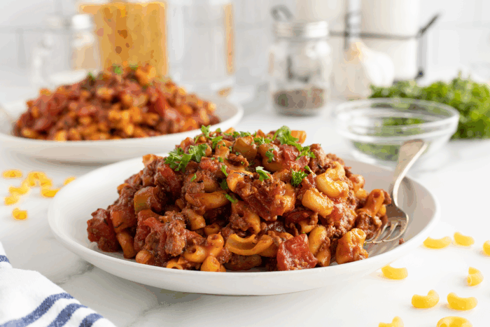 A plate of macaroni pasta with ground beef, tomato sauce, and chopped parsley sits on a white table with a fork. Another plate, glass jars, and fresh herbs are visible in the background. Uncooked pasta is scattered nearby.