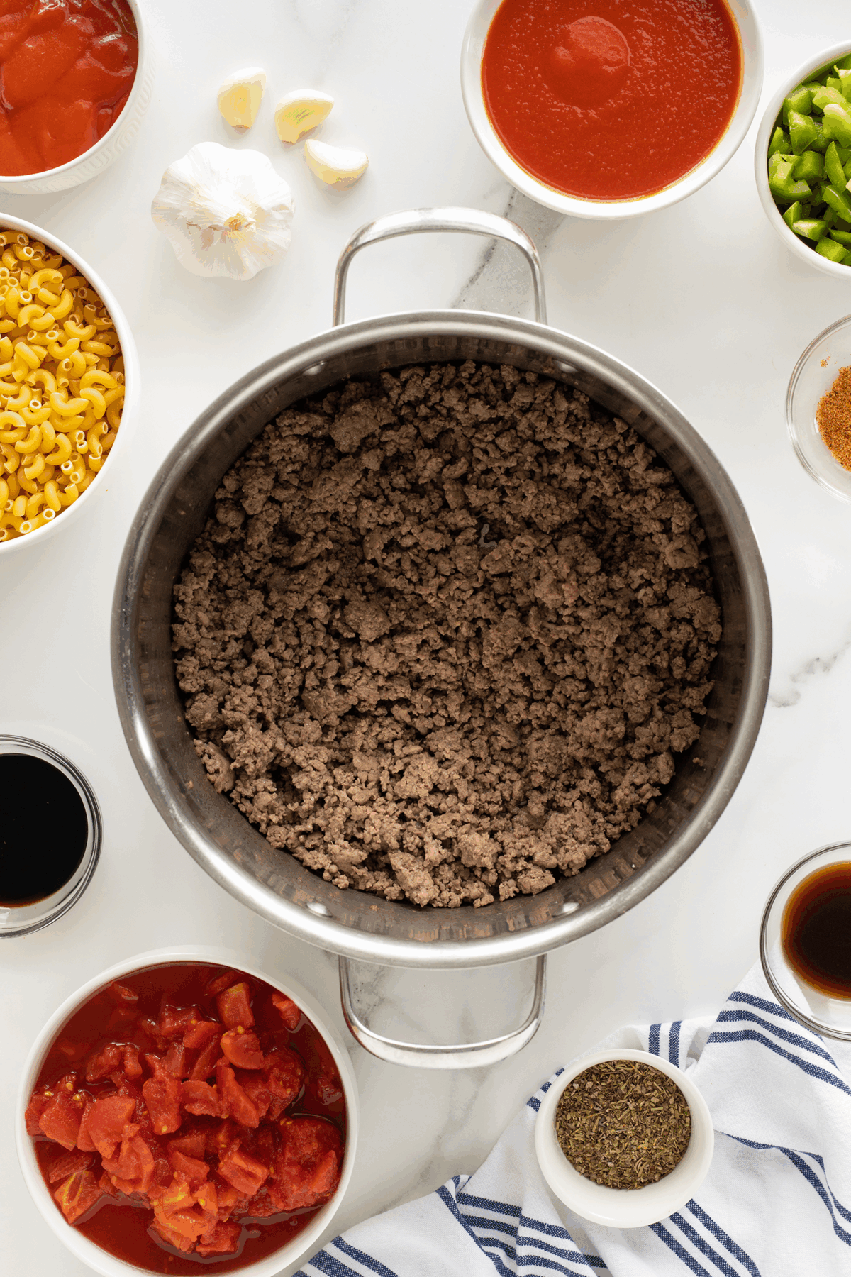 Overhead view of a pot filled with cooked ground beef, surrounded by small bowls of pasta, tomatoes, tomato sauce, seasonings, green peppers, garlic cloves, and liquids on a white marble surface.