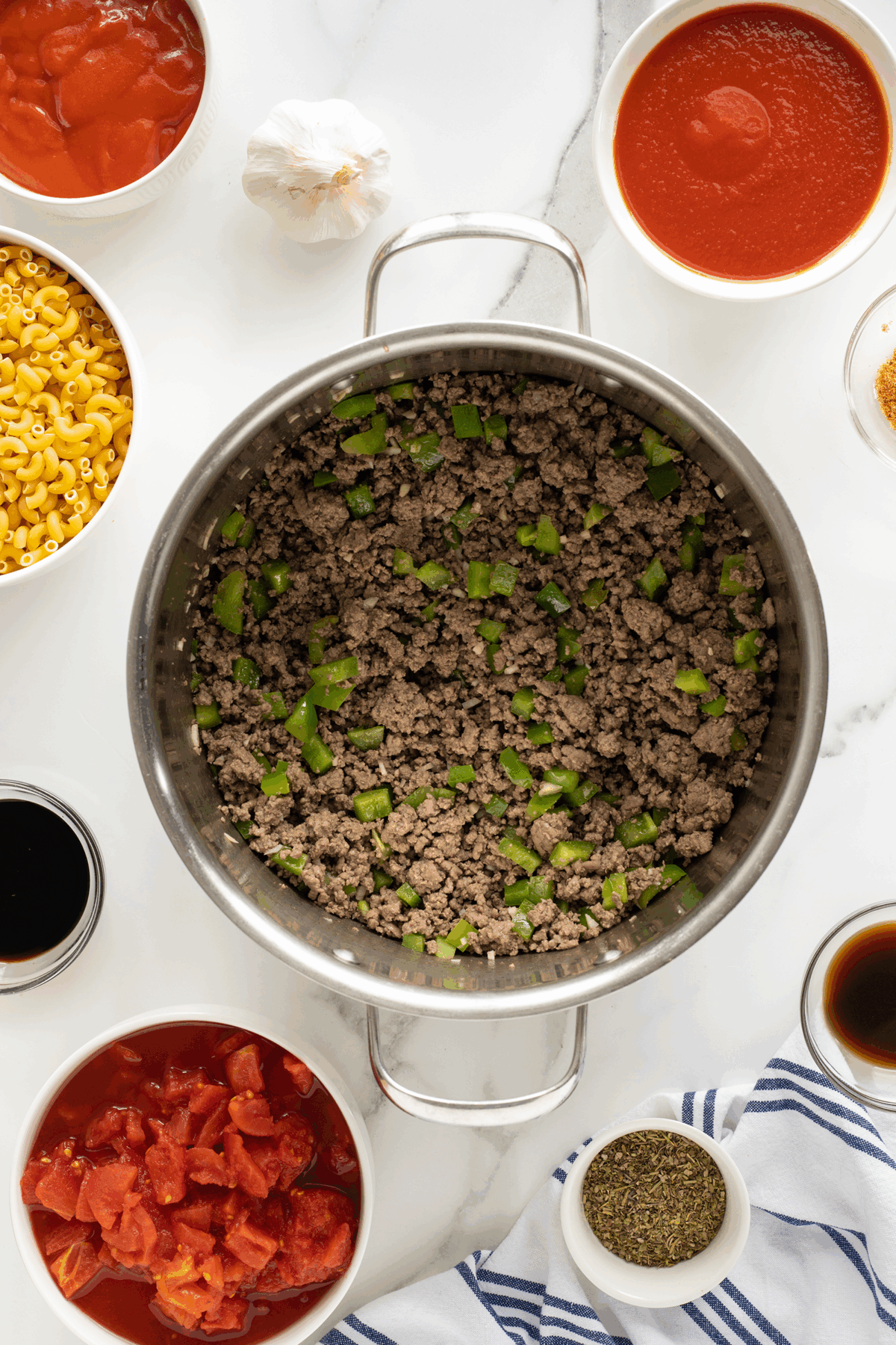 A pot of cooked ground beef and chopped green peppers sits on a white countertop, surrounded by bowls of uncooked pasta, tomato sauce, canned tomatoes, garlic, seasonings, and sauces.