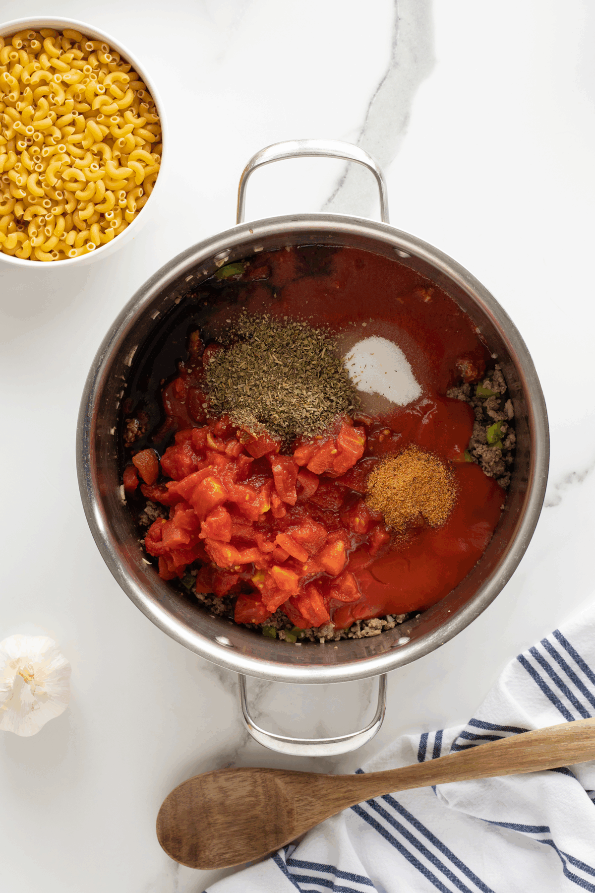 A pot on a white countertop contains ground meat, diced tomatoes, tomato sauce, Italian herbs, sugar, and seasoning. Nearby are a bowl of dry elbow macaroni, a garlic bulb, a wooden spoon, and a striped towel.
