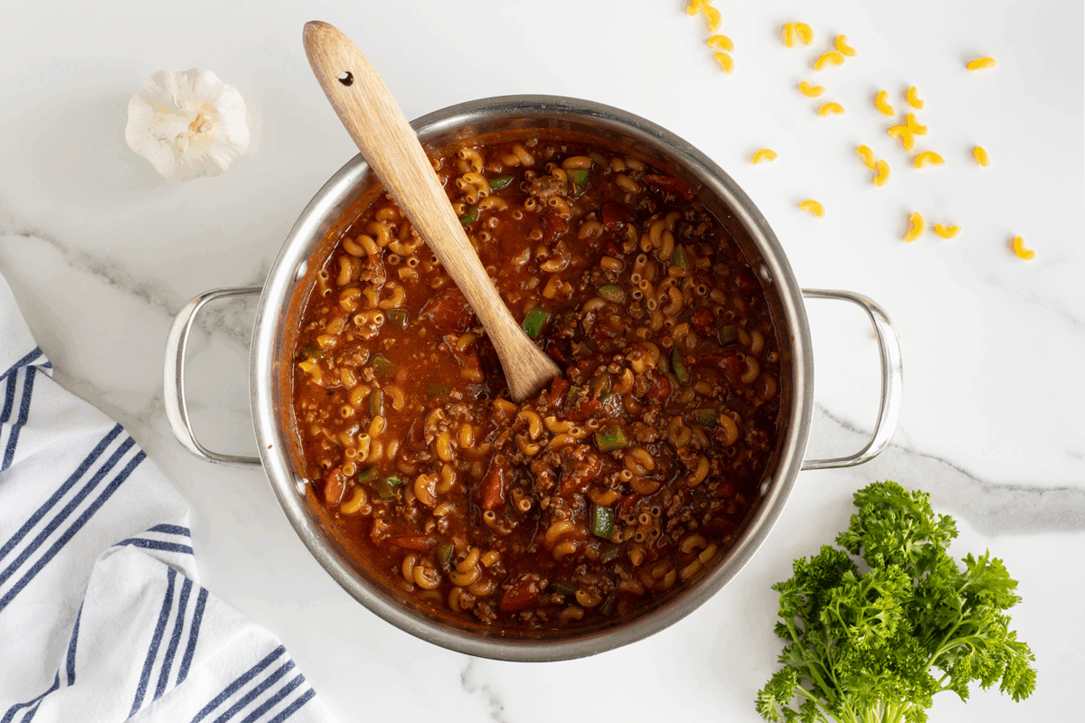 A pot of tomato-based macaroni soup with vegetables and a wooden spoon sits on a white marble surface, surrounded by a head of garlic, scattered macaroni, a bunch of parsley, and a striped kitchen towel.