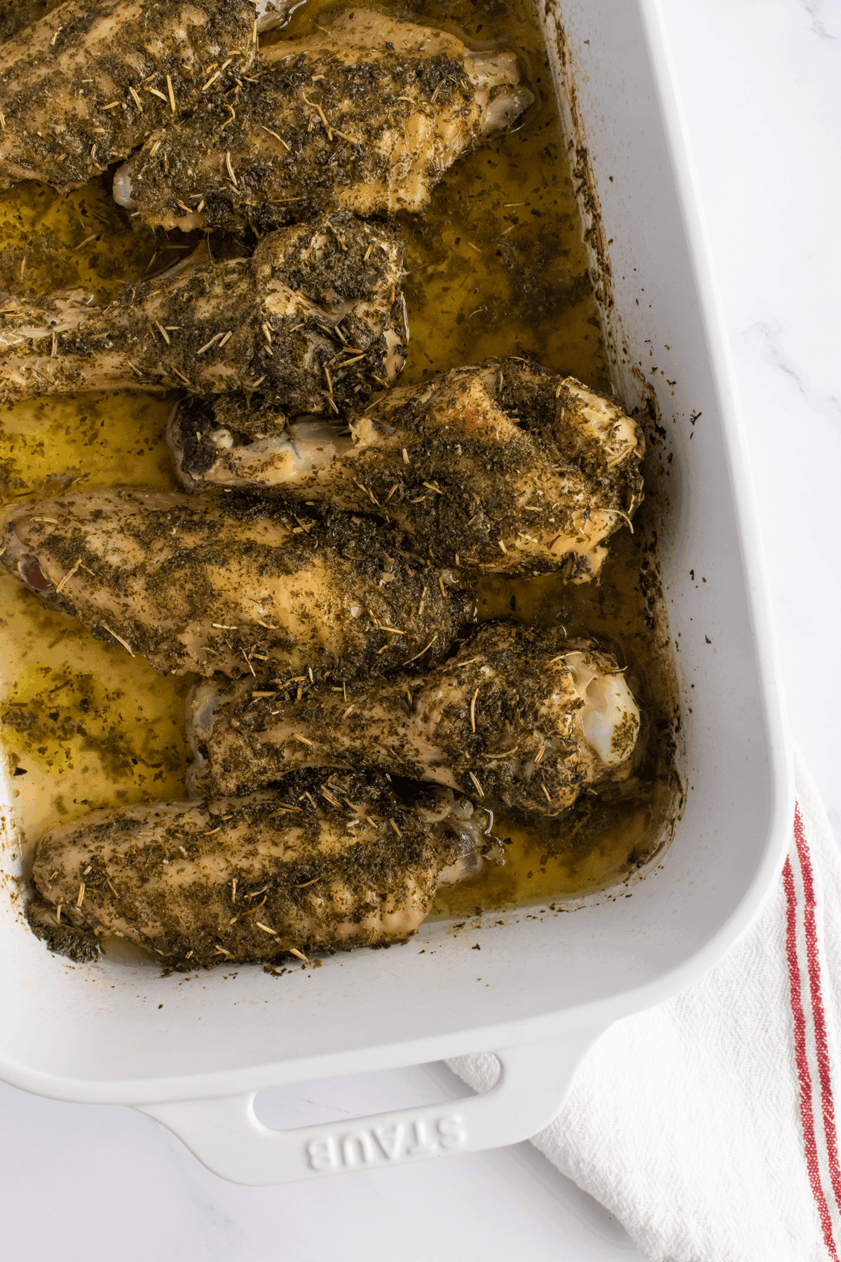 Baked turkey wings coated with herbs and spices in a white rectangular baking dish, sitting on a white surface with a white and red striped kitchen towel beside it.