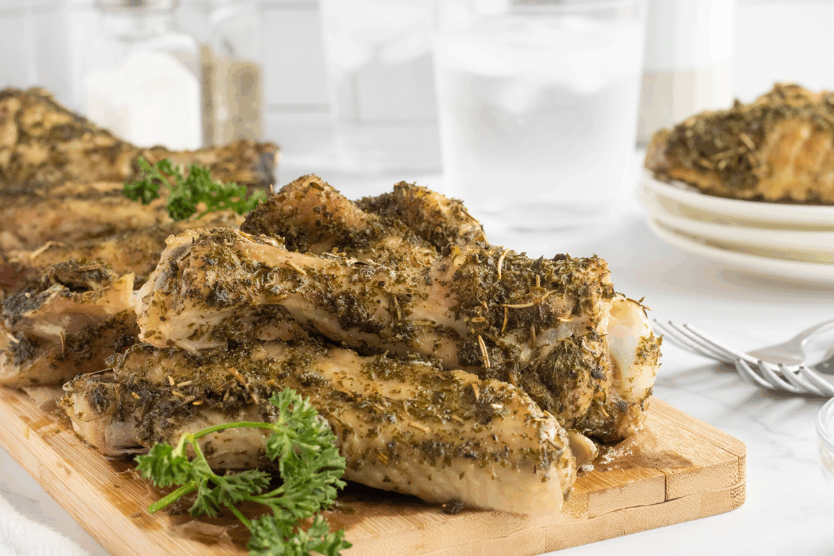 A pile of seasoned, cooked pork ribs garnished with parsley is arranged on a wooden cutting board. Plates of food and glasses of ice water are in the background on a white table.