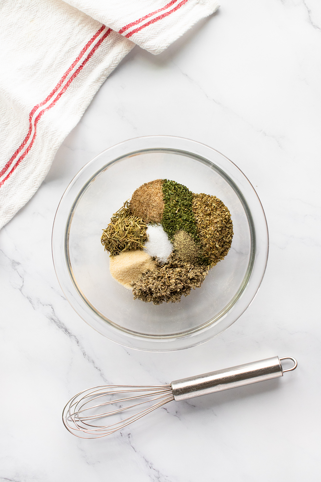 A glass bowl filled with assorted dried herbs and spices sits on a marble surface next to a metal whisk; a white towel with red stripes is in the upper left corner.