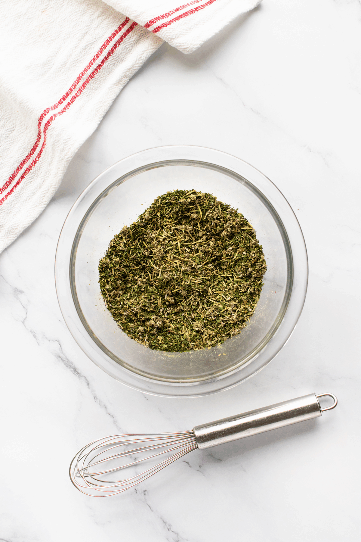 A glass bowl filled with dried mixed herbs sits on a white marble surface next to a metal whisk. A white kitchen towel with red stripes is in the corner of the image.