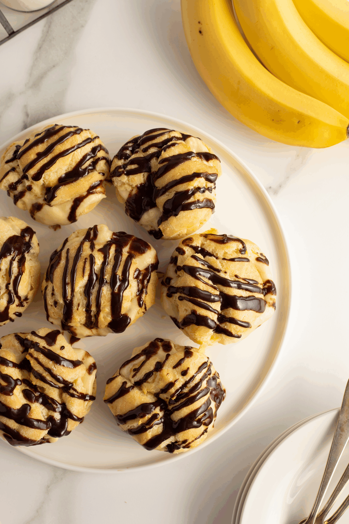 A plate of six cream puffs drizzled with chocolate sauce sits on a white surface next to a bunch of bananas and a stack of white dishes with silver utensils.