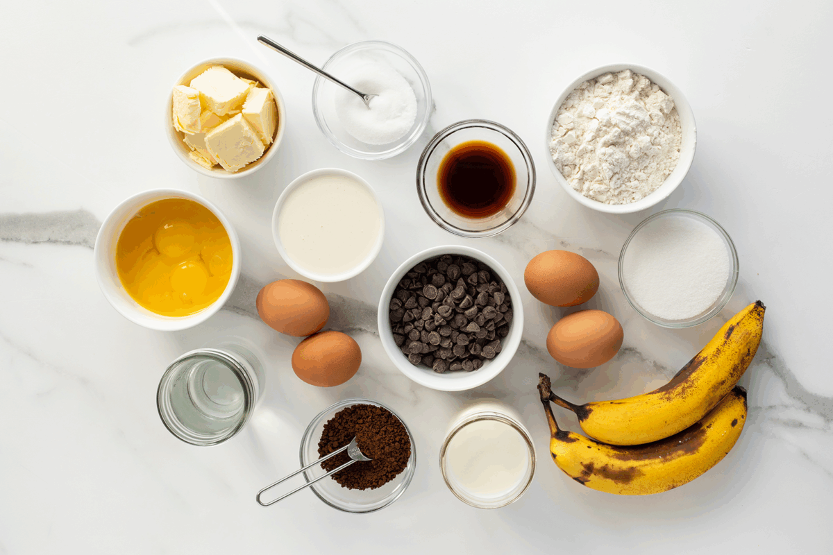 Assorted baking ingredients on a white surface, including butter, sugar, eggs, cream, chocolate chips, vanilla, flour, bananas, milk, coffee grounds, and water, each in separate bowls or containers.