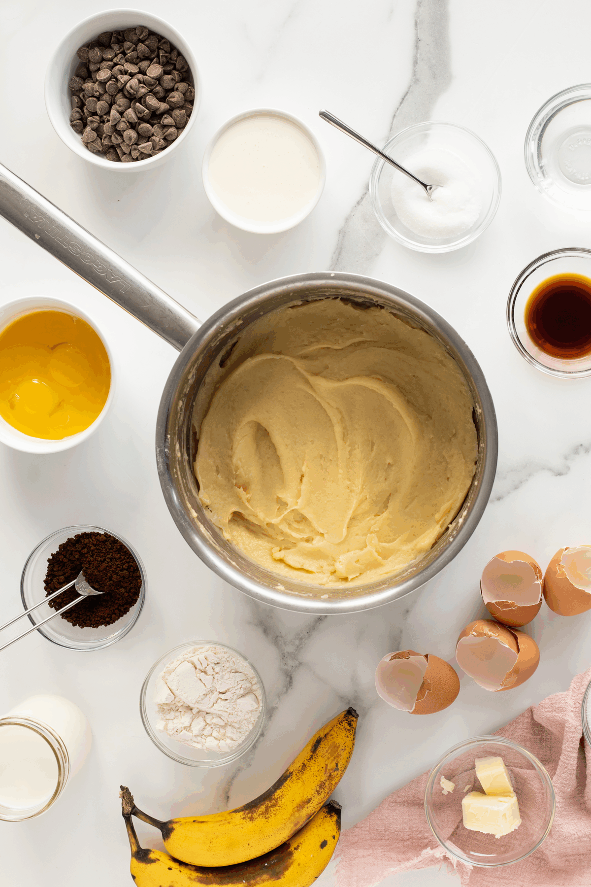 A saucepan of creamy batter is surrounded by baking ingredients on a white surface: chocolate chips, cream, sugar, vanilla, egg yolks, coffee, flour, eggshells, milk, ripe bananas, and butter.