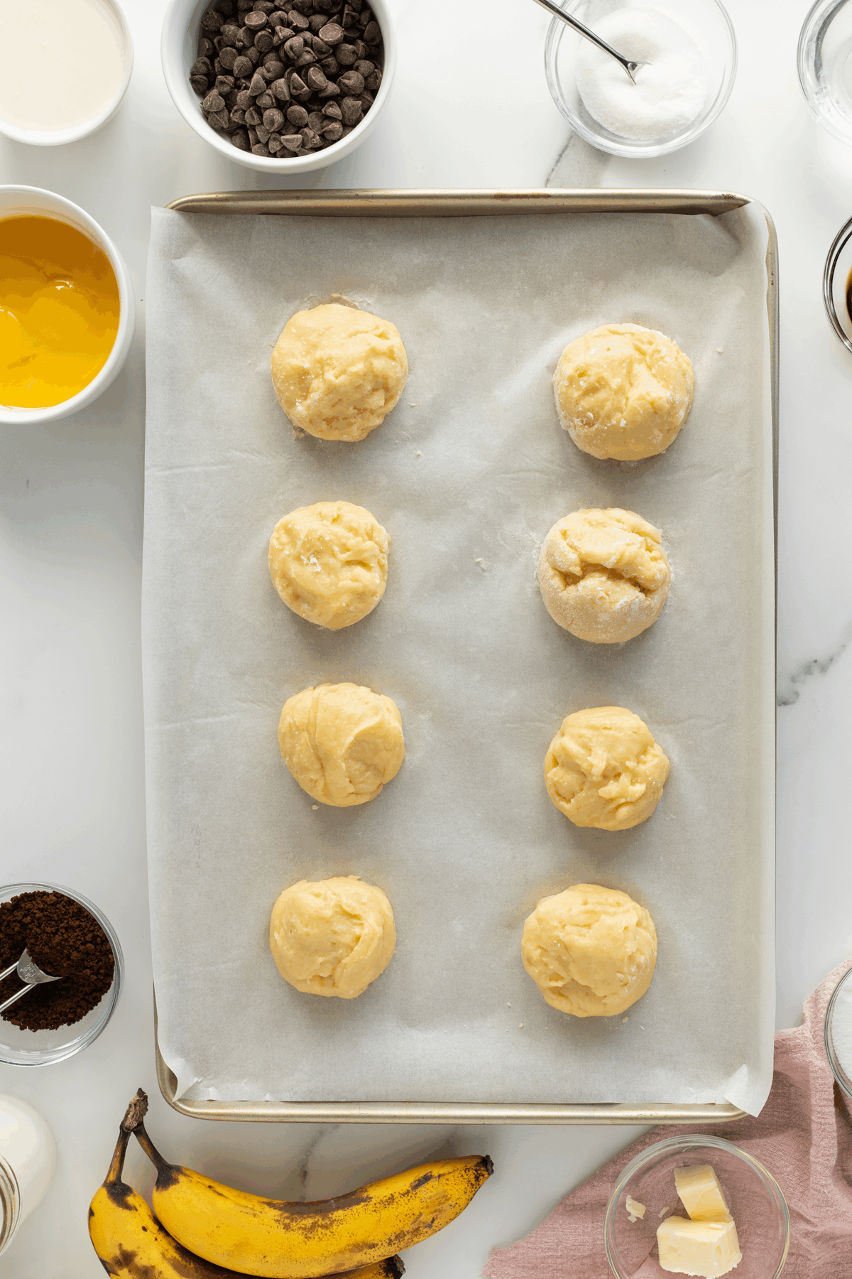 A baking tray lined with parchment paper holds eight unbaked cookie dough balls. Surrounding the tray are ingredients in bowls, including eggs, chocolate chips, sugar, butter, cream, instant coffee, and a ripe banana.