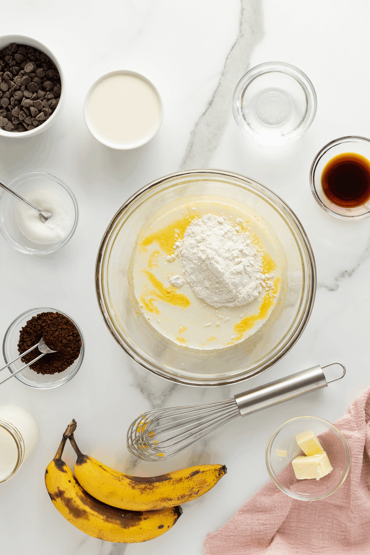 A top view of baking ingredients on a white surface, including flour, sugar, butter, chocolate chips, vanilla, cream, yogurt, coffee grounds, two ripe bananas, a whisk, and a pink cloth.