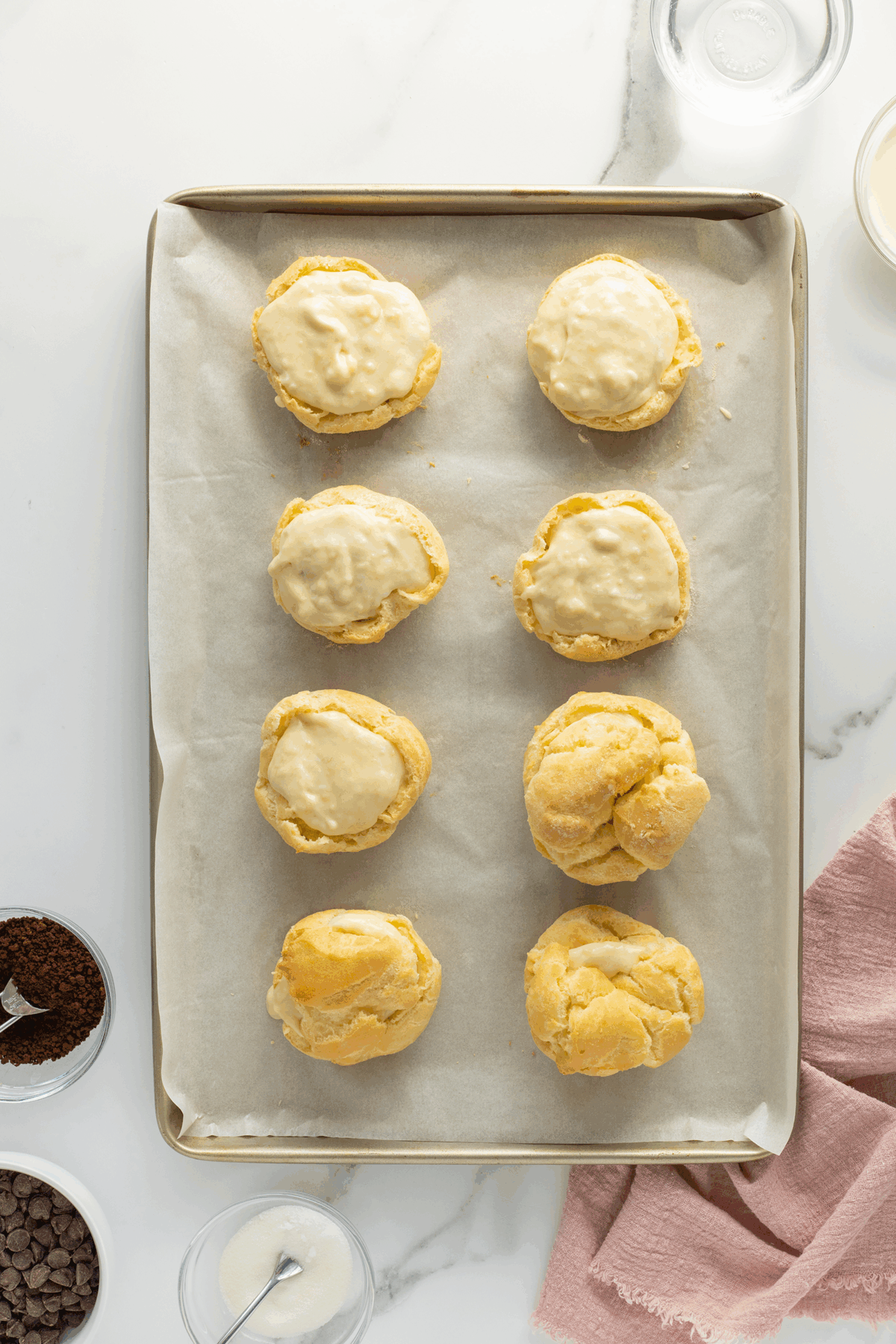 A baking tray lined with parchment paper holds eight golden cream puffs, some glazed, on a marble counter. Surrounding the tray are small bowls with ingredients and a pink cloth napkin.