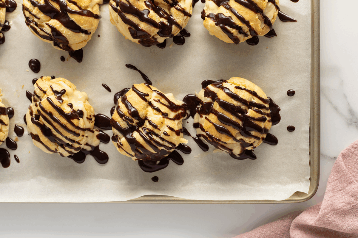 A baking tray with cream puffs filled with custard, drizzled generously with chocolate sauce, lined up on parchment paper. A pink napkin is partially visible in the corner of the image.