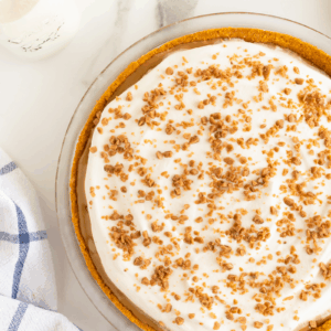 A cream pie with a crumbly crust, topped with whipped cream and sprinkled toffee bits, sits on a marble surface next to a milk bottle, a blue-and-white towel, and a small bowl of toffee bits.
