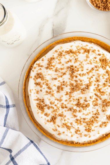 A cream pie with a crumbly crust, topped with whipped cream and sprinkled toffee bits, sits on a marble surface next to a milk bottle, a blue-and-white towel, and a small bowl of toffee bits.