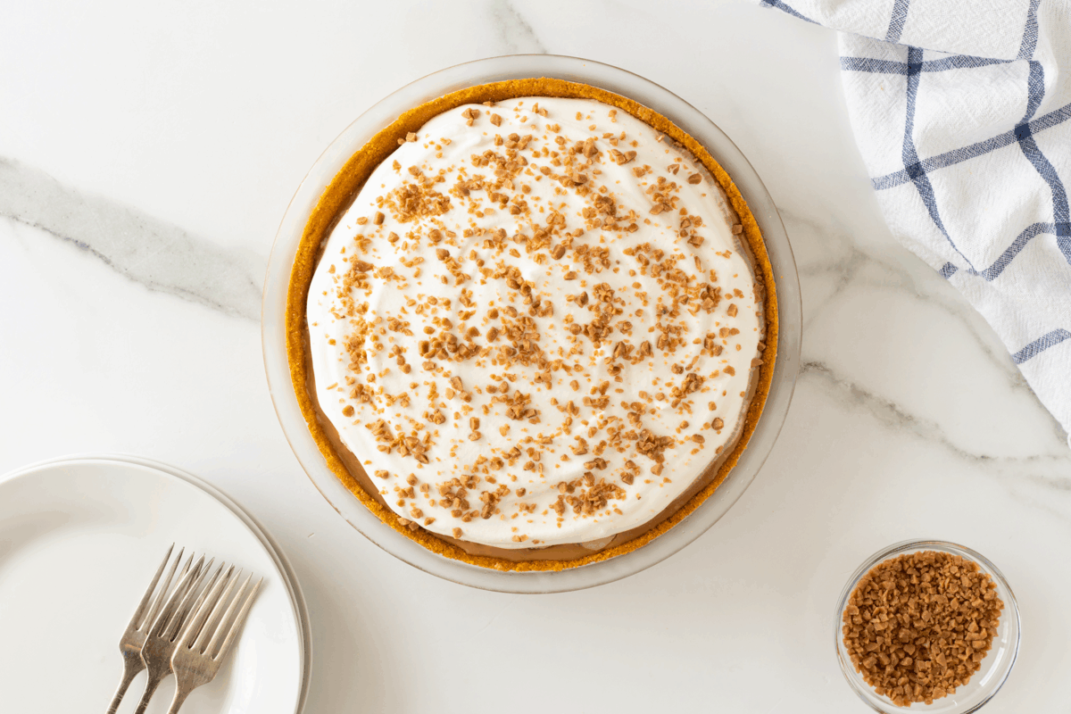 A round pie with a creamy topping and brown crumbly sprinkles sits on a white marble surface next to a plate with two forks, a bowl of crumbs, and a white towel with blue stripes.