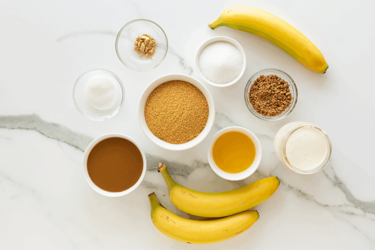 Overhead view of three bananas and small bowls containing brown sugar, granulated sugar, honey, ginger, milk, peanut butter, and a brown powder, all arranged on a white marble surface.