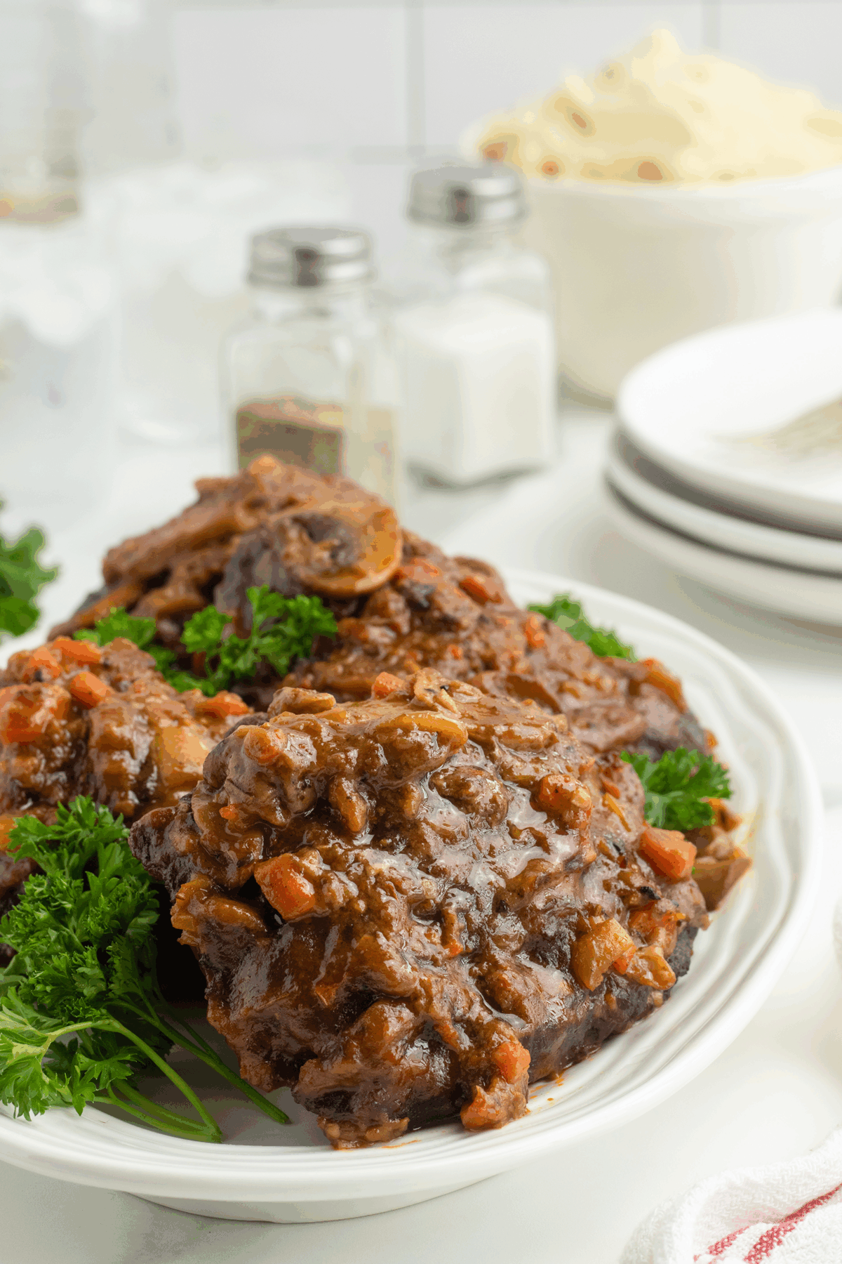 A plate of braised oxtails in a rich, brown sauce with carrots and onions, garnished with fresh parsley. In the background are plates, utensils, a bowl of mashed potatoes, and salt and pepper shakers.