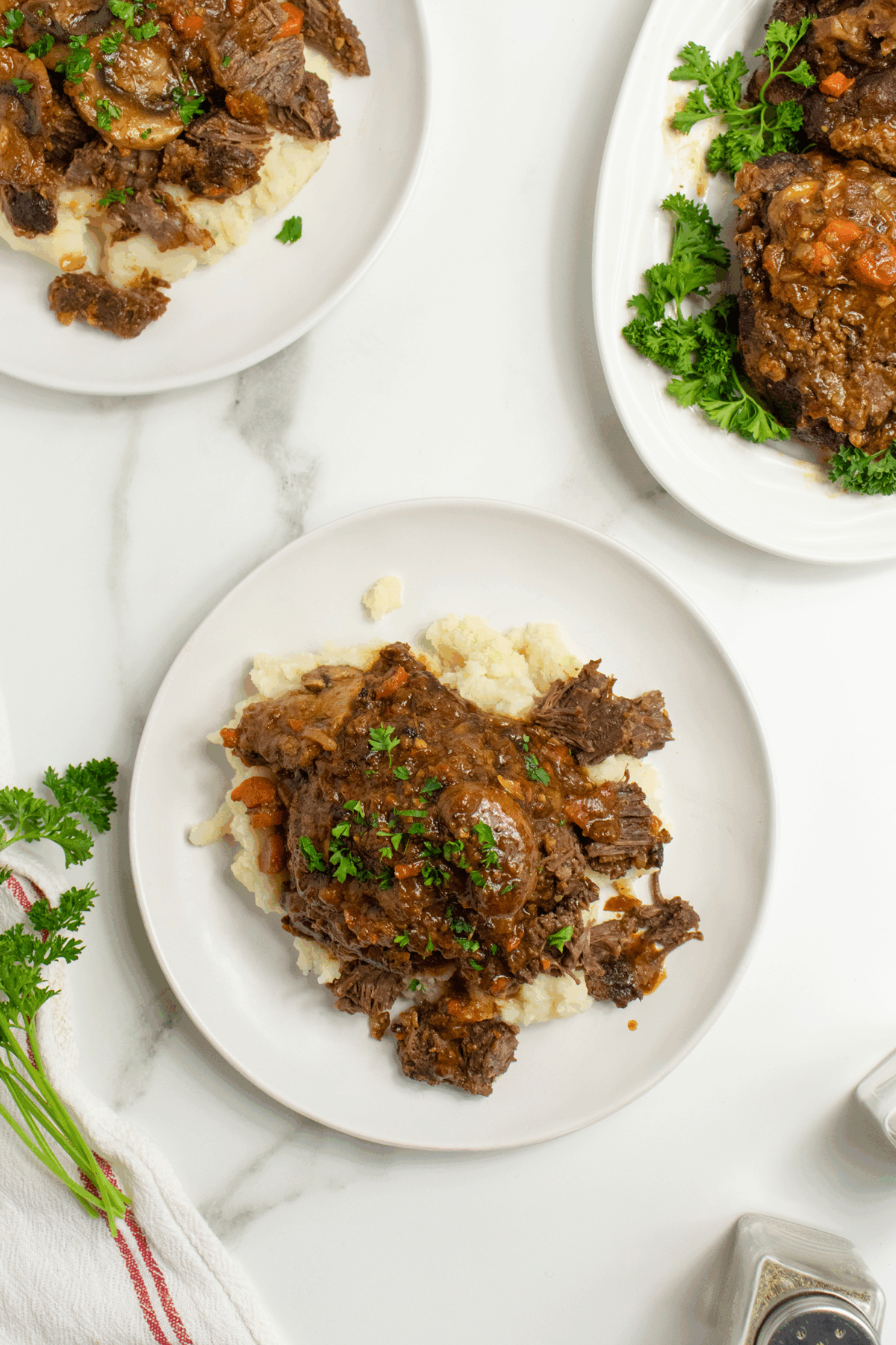 Three white plates with servings of braised meat in sauce over mashed potatoes, garnished with chopped parsley, on a white marble surface with a sprig of parsley and a striped kitchen towel nearby.