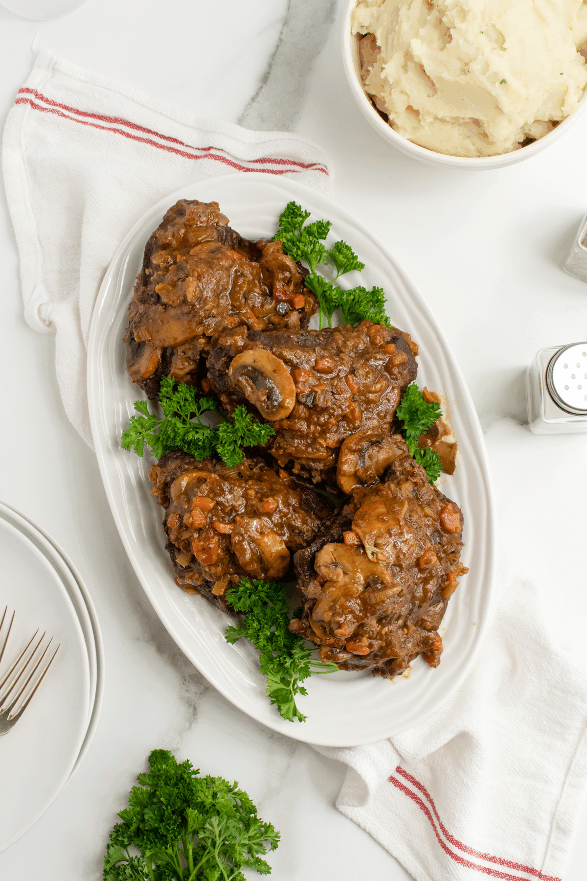 A white oval platter with braised beef short ribs topped with vegetables and garnished with parsley, placed on a white surface next to a bowl of mashed potatoes and a white napkin with a red stripe.