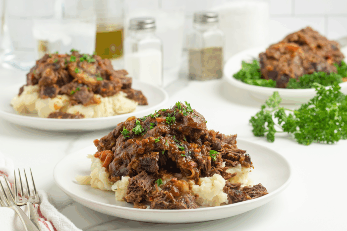 Plates of shredded pot roast with gravy served over mashed potatoes, garnished with chopped parsley. Fresh parsley and seasonings are in the background, along with glasses of water and a fork on the table.