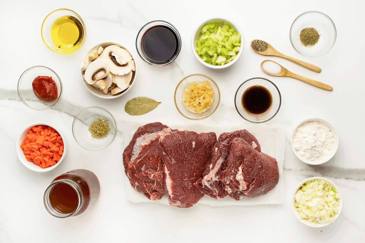 Raw beef, surrounded by bowls of chopped carrots, celery, onions, mushrooms, minced garlic, flour, tomato paste, broth, red wine, oil, Worcestershire sauce, and spices, all arranged on a white surface.