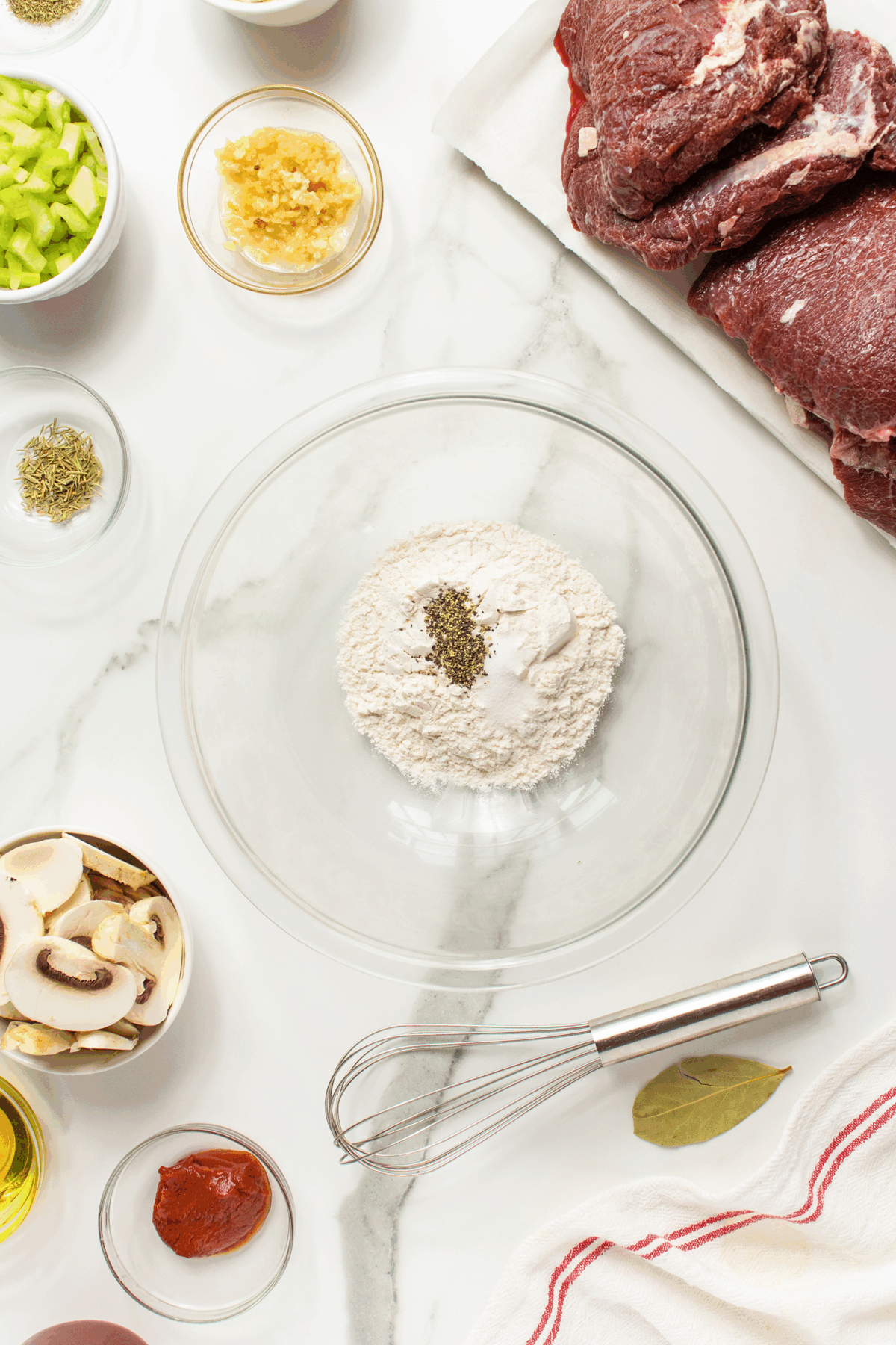 A glass bowl with flour and herbs sits on a marble counter surrounded by raw beef, mushrooms, chopped celery, minced garlic, seasoning, tomato paste, oil, a whisk, and a bay leaf.