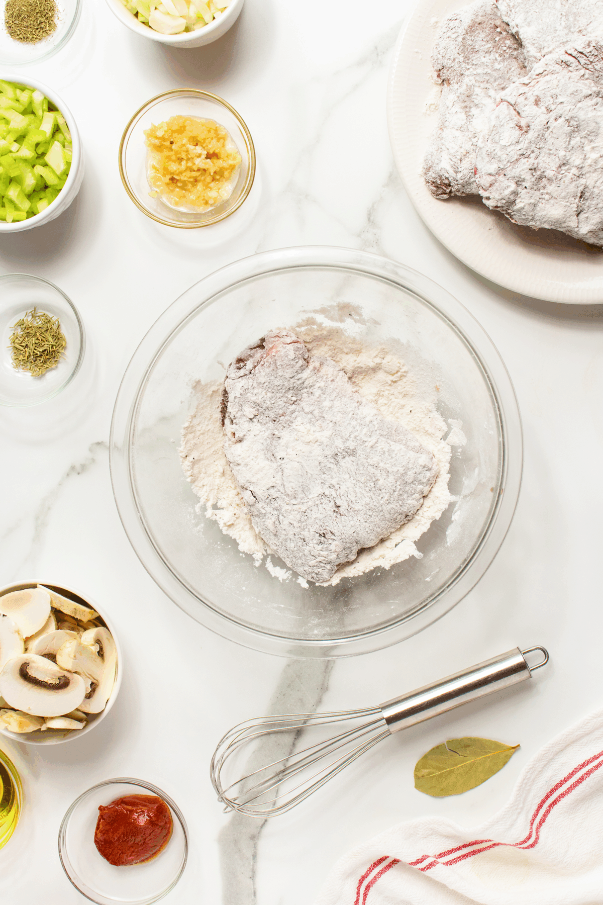 A glass bowl with a flour-coated piece of meat, surrounded by bowls of chopped celery, mushrooms, herbs, minced garlic, tomato paste, and a whisk on a marble surface.