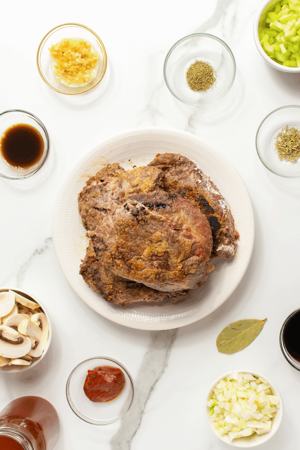 A white plate with cooked beef sits in the center, surrounded by small bowls of chopped onion, celery, mushrooms, tomato paste, minced garlic, dried herbs, bay leaf, sauces, and seasonings on a white countertop.
