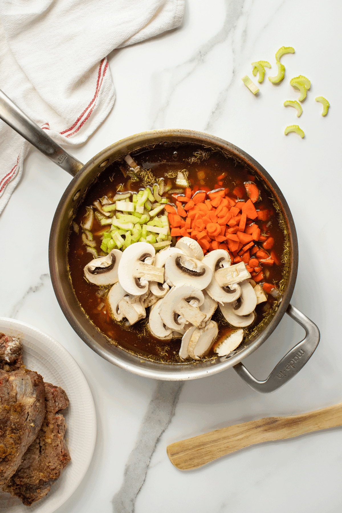 A saucepan filled with broth, sliced mushrooms, chopped celery, and diced carrots sits on a white marble countertop. Next to it are a wooden spoon, a white towel, a few celery slices, and a plate with cooked meat.