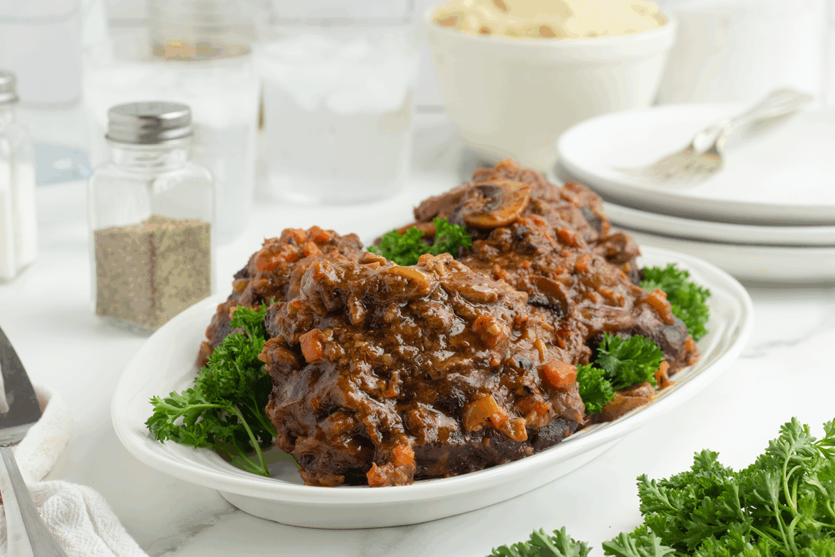 A white platter holds braised oxtails in a rich, tomato-based sauce, garnished with fresh parsley. In the background are plates, forks, a bowl of mashed potatoes, salt and pepper shakers, and glasses of iced water.