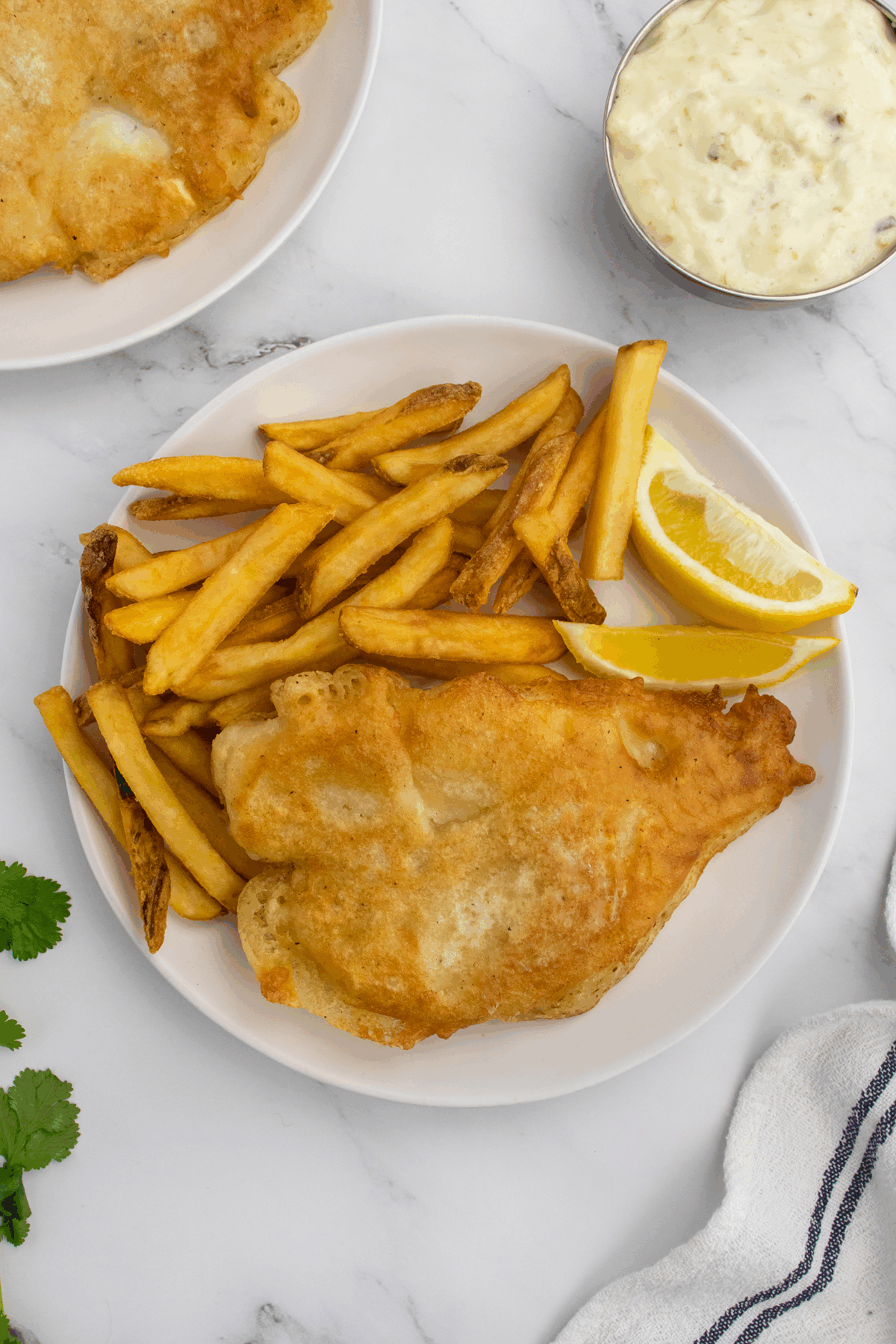 A plate with golden battered fish, French fries, and a lemon wedge on a white surface. A bowl of tartar sauce and part of another plate are visible nearby.