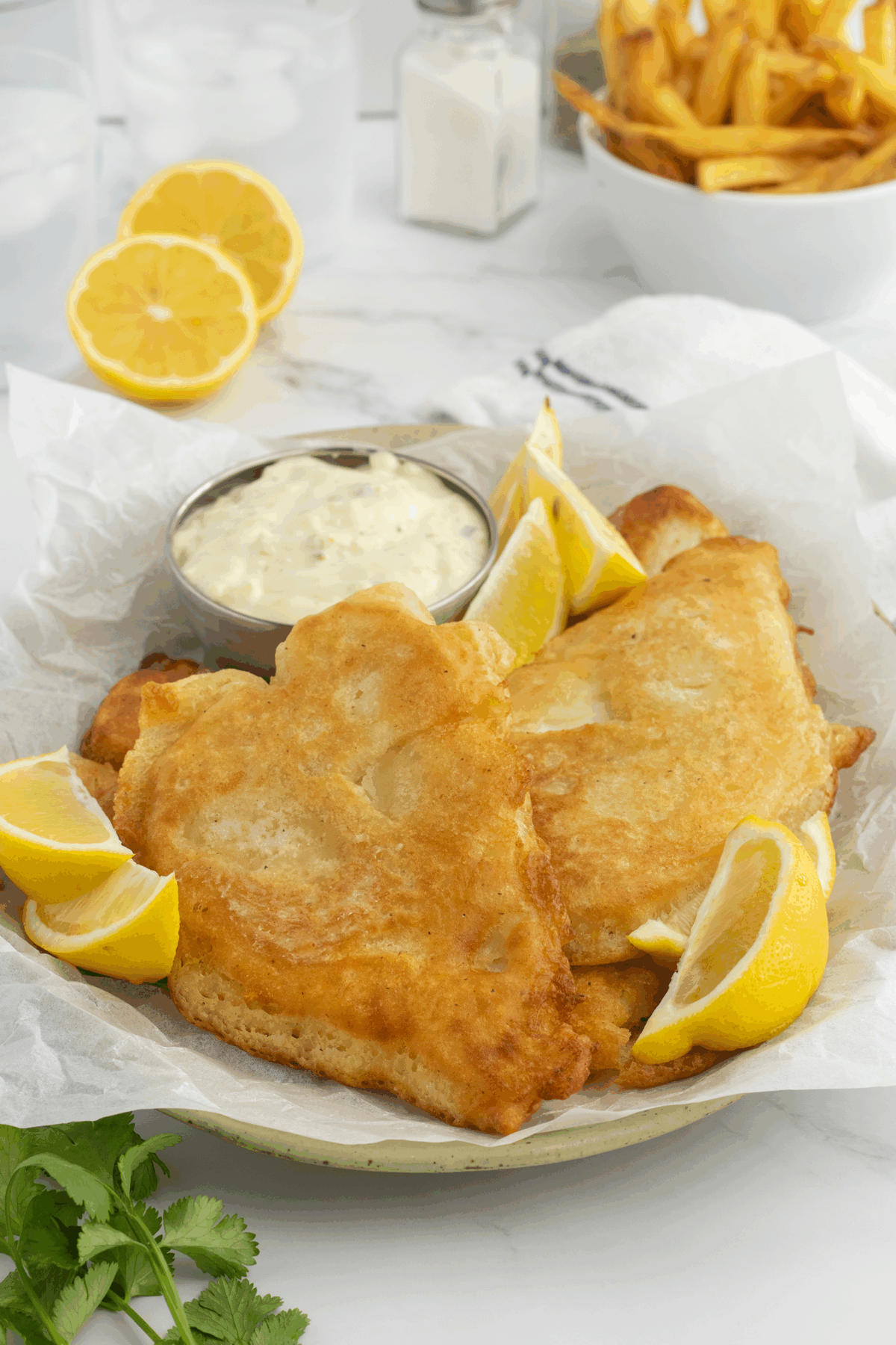Two pieces of golden fried fish with lemon wedges and a side of tartar sauce on parchment paper, with fries, lemon halves, and drinks in the background.